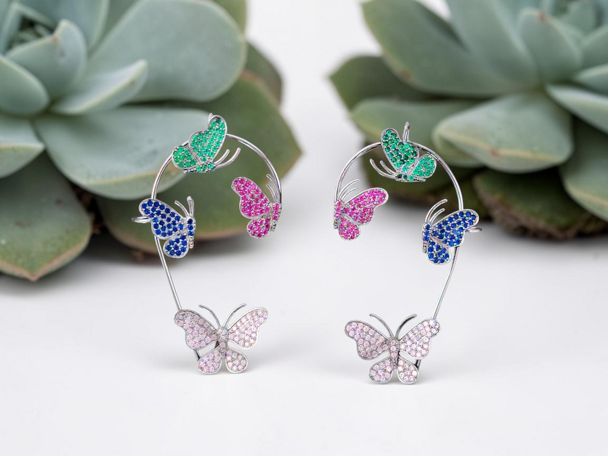 A pair of silver-toned hoop earrings decorated with multiple butterfly charms in green, blue, pink, and lavender, displayed against a white background with blurred green succulents.