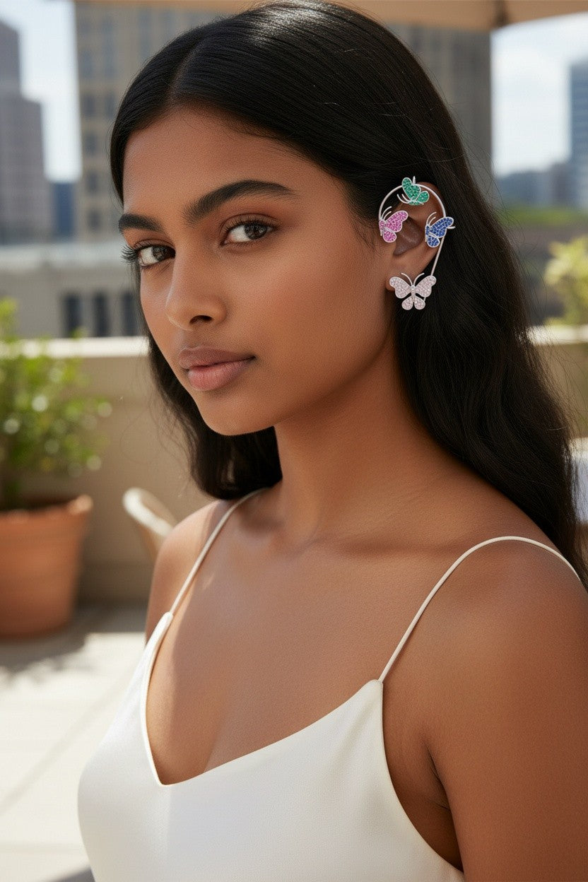 A woman with long dark hair wearing a white spaghetti-strap top and a colorful butterfly-shaped ear cuff, photographed outdoors on a balcony with a blurred city skyline in the background.