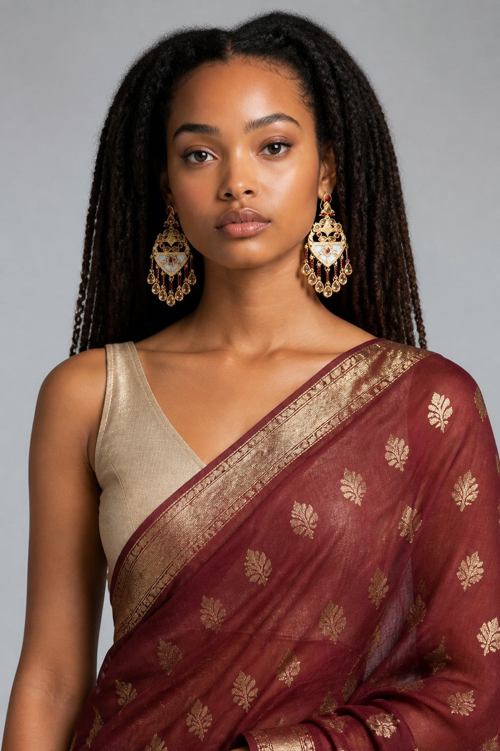 A woman wearing a deep maroon silk saree with gold floral motifs and a gold border, paired with a beige sleeveless blouse and large gold earrings, against a neutral gray background.