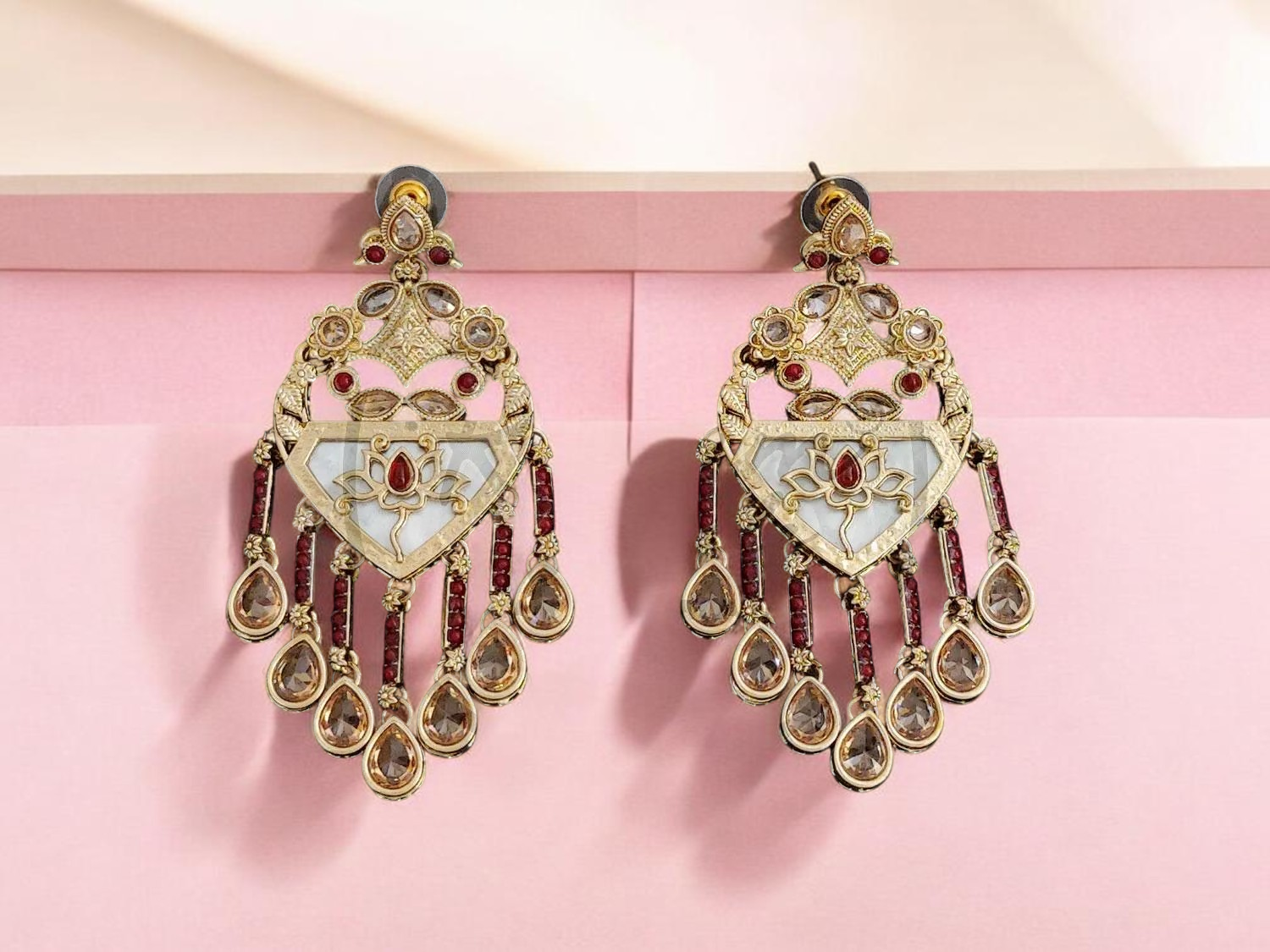 A pair of ornate gold-toned statement earrings with white mother-of-pearl inlay, small red gemstone accents, and dangling teardrop charms, displayed against a soft pink background.